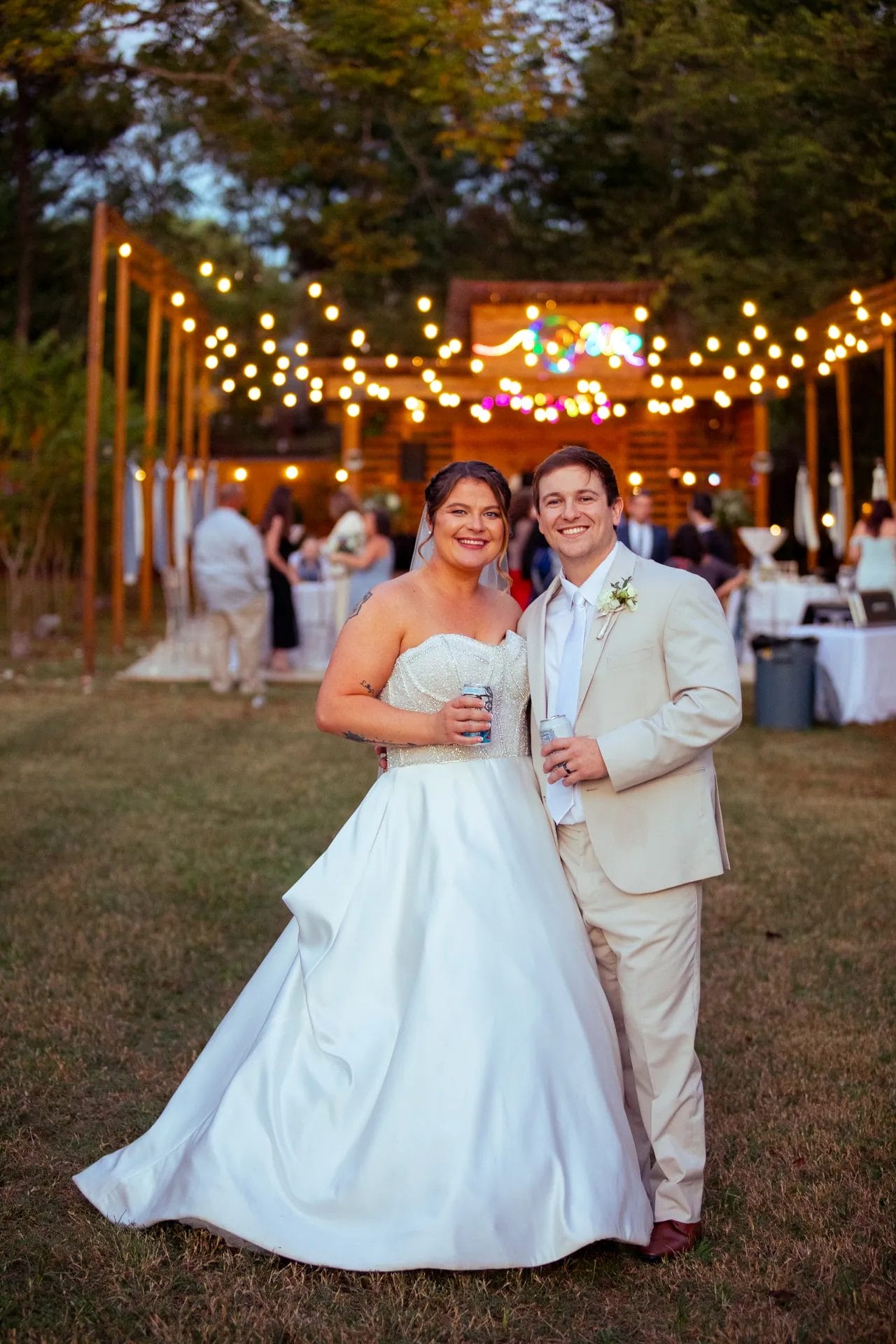 A bride and groom smile at an outdoor wedding reception. The bride wears a white gown, and the groom wears a beige suit. They hold drinks, standing on grass under string lights, with guests and tables in the background. Huntersville event venue