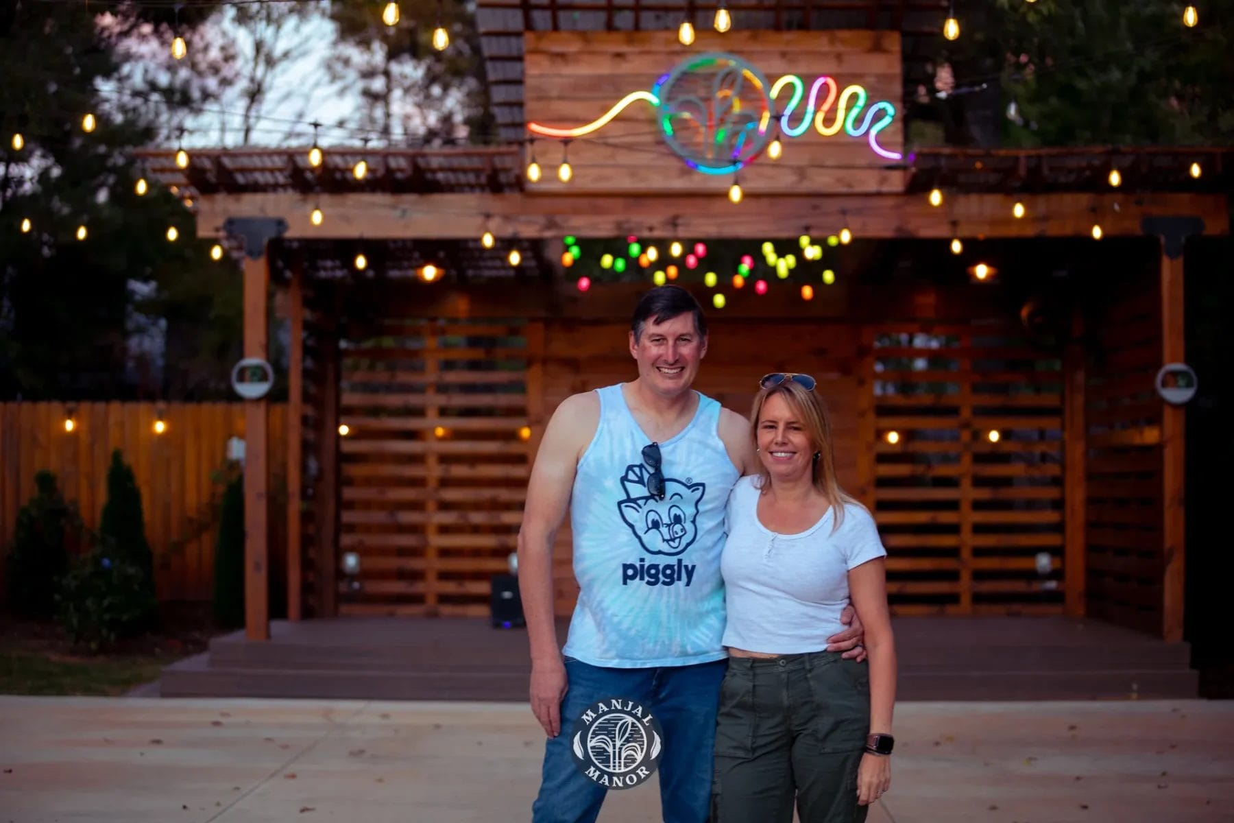 A man and woman stand smiling in front of a wooden structure with colorful string lights and a neon sign above. The man wears a "piggly" tank top, while the woman wears a white t-shirt. The scene has a relaxed and cheerful ambiance. Huntersville event venue