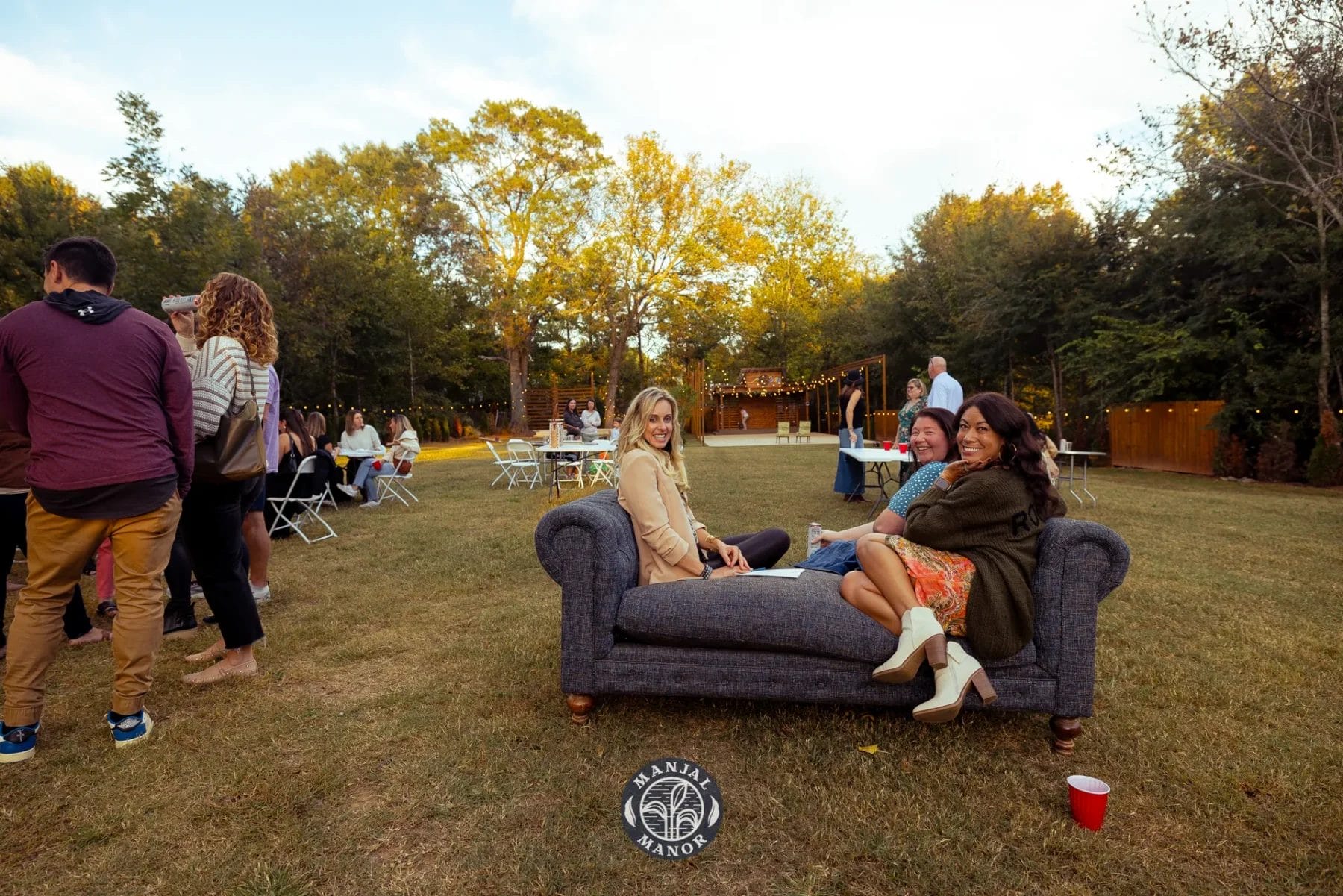 Two women sitting on a couch outdoors, smiling at the camera. People are standing around in the background at an outdoor gathering. The setting is a grassy area with trees, and the sun is low in the sky. Tables and chairs are set up nearby. Huntersville event venue