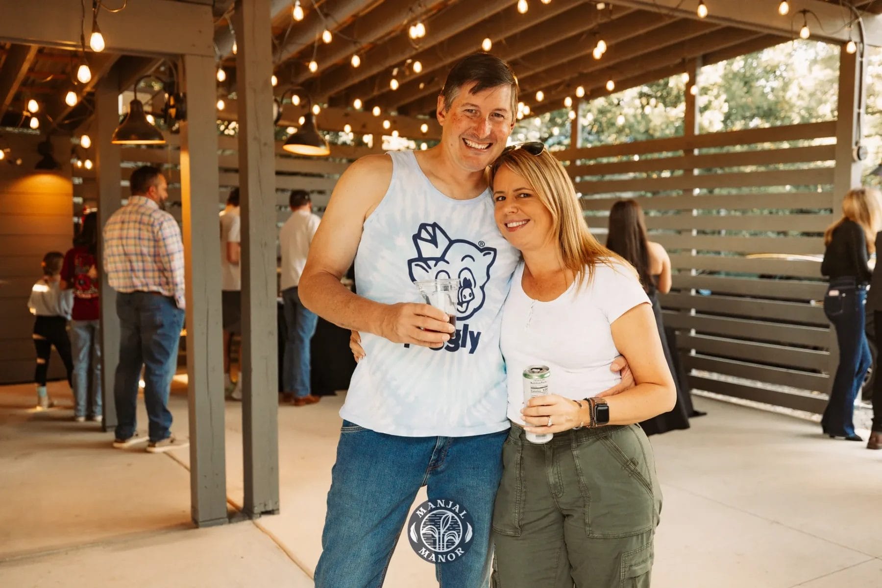 A man and woman are smiling at an outdoor gathering. The man is wearing a tank top with a bear design, and the woman is in a white t-shirt and green pants. They are each holding a drink. String lights and people are visible in the background. Huntersville event venue