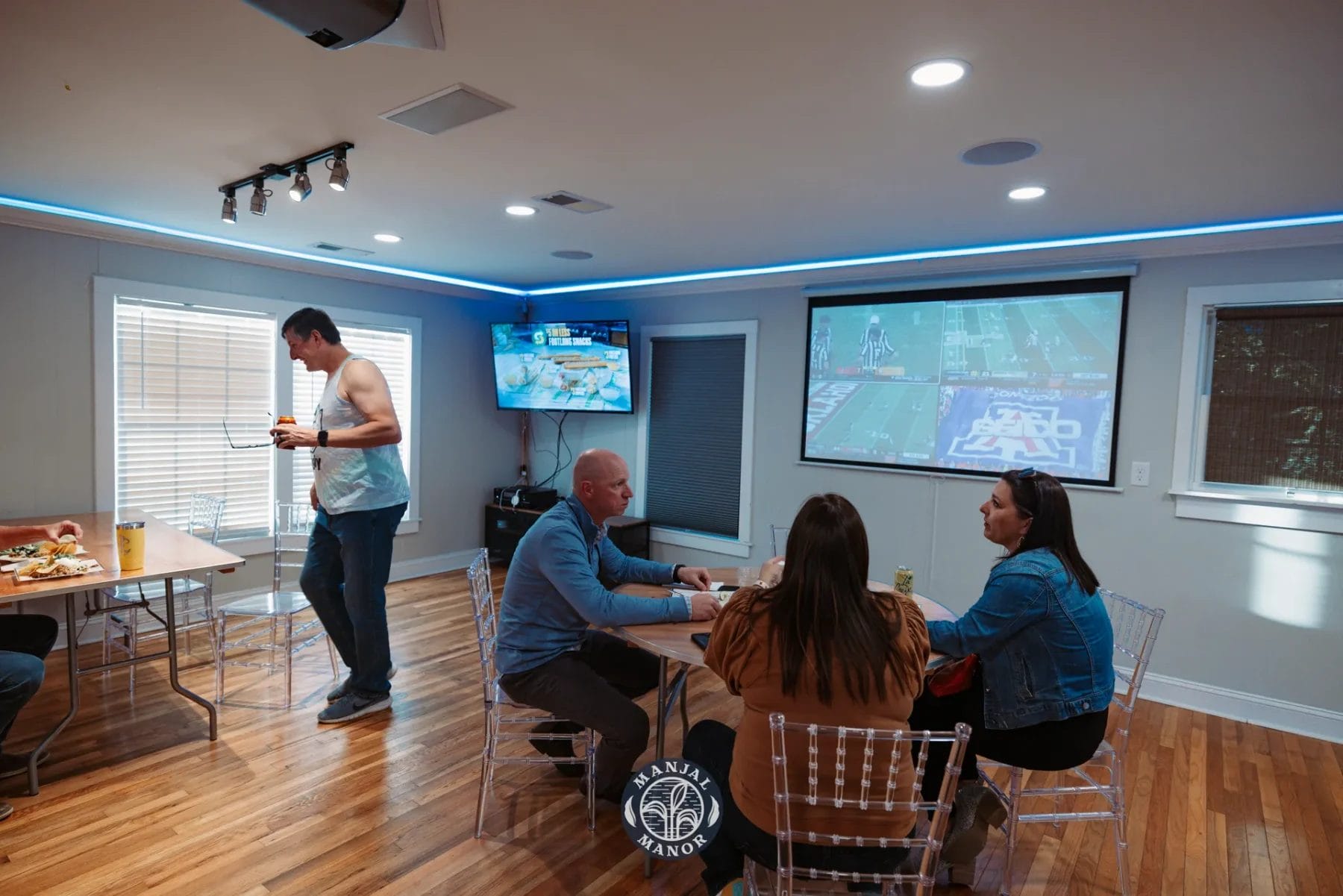 People are sitting and eating in a room with a wooden floor. There are multiple large screens on the walls showing a football game. One person is standing by a table with food and drinks. The room is softly lit with blue accent lights. Huntersville event venue