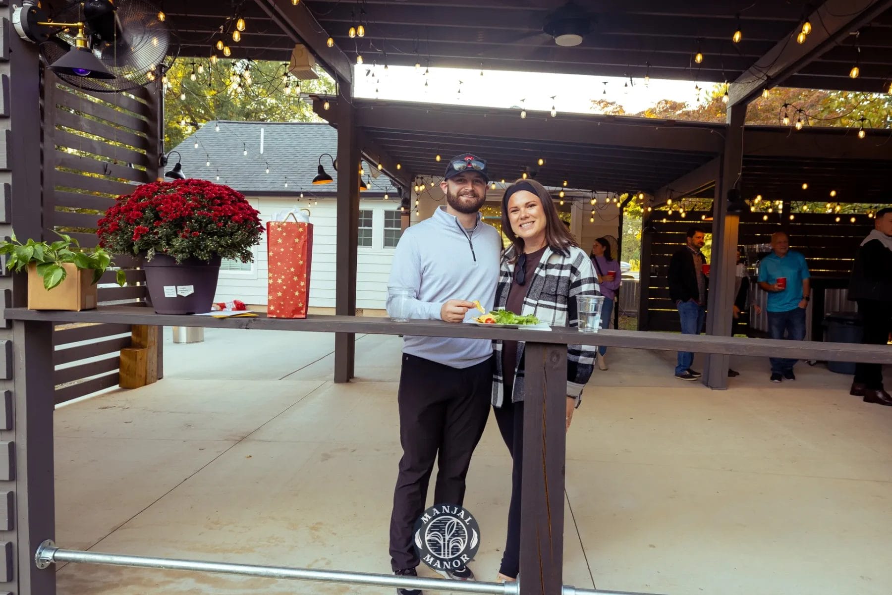 A smiling couple stands under a pergola adorned with string lights. They hold drinks and food on a ledge. Decorated with plants and a gift bag, the outdoor space has a festive atmosphere, with other people visible in the background. Huntersville event venue