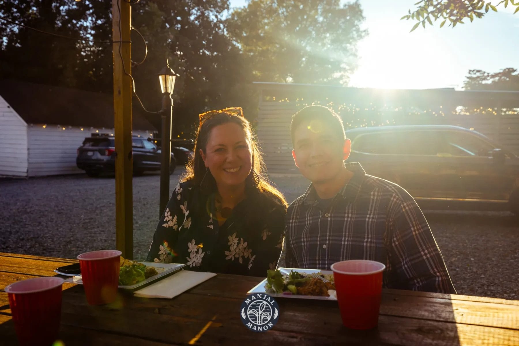 A woman and a man smile while seated at a wooden picnic table with red cups and plates of food. The sun is shining brightly, creating a warm glow. In the background, there are parked cars and a white building. Huntersville event venue