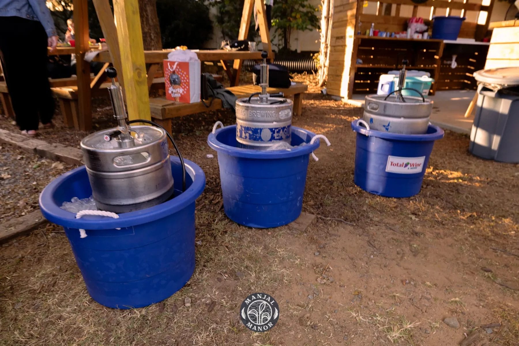 Three metal beer kegs chilled in blue plastic tubs filled with ice on the ground, outdoors. The scene is set near a wooden structure with various party supplies scattered in the background. Huntersville event venue