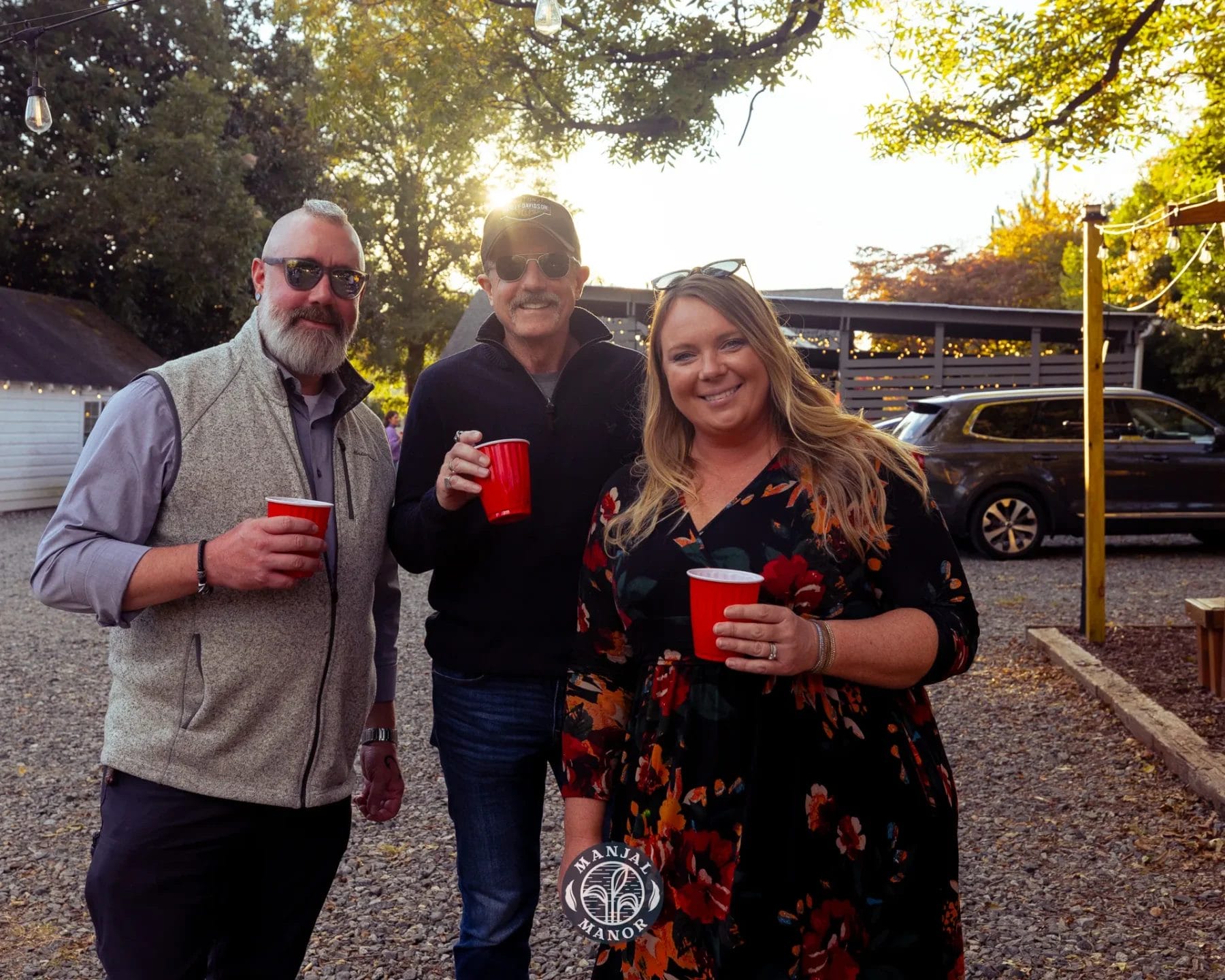 Three people stand outdoors, each holding a red cup. They are smiling and dressed casually, with trees and parked cars in the background. The sun is setting, casting a warm glow over the scene. Huntersville event venue