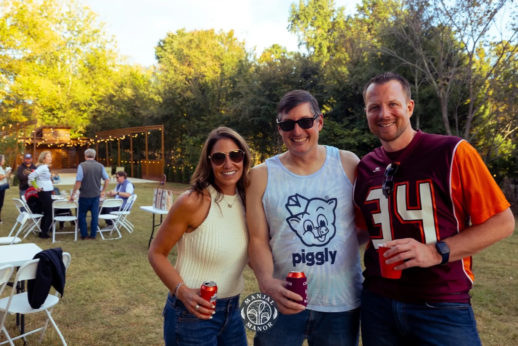 Three people smiling at an outdoor gathering. The woman wears sunglasses and a light top, the middle man sports a "piggly" tank top, and the man on the right wears a burgundy jersey. They hold drinks, and picnic tables are visible in the background. Huntersville event venue