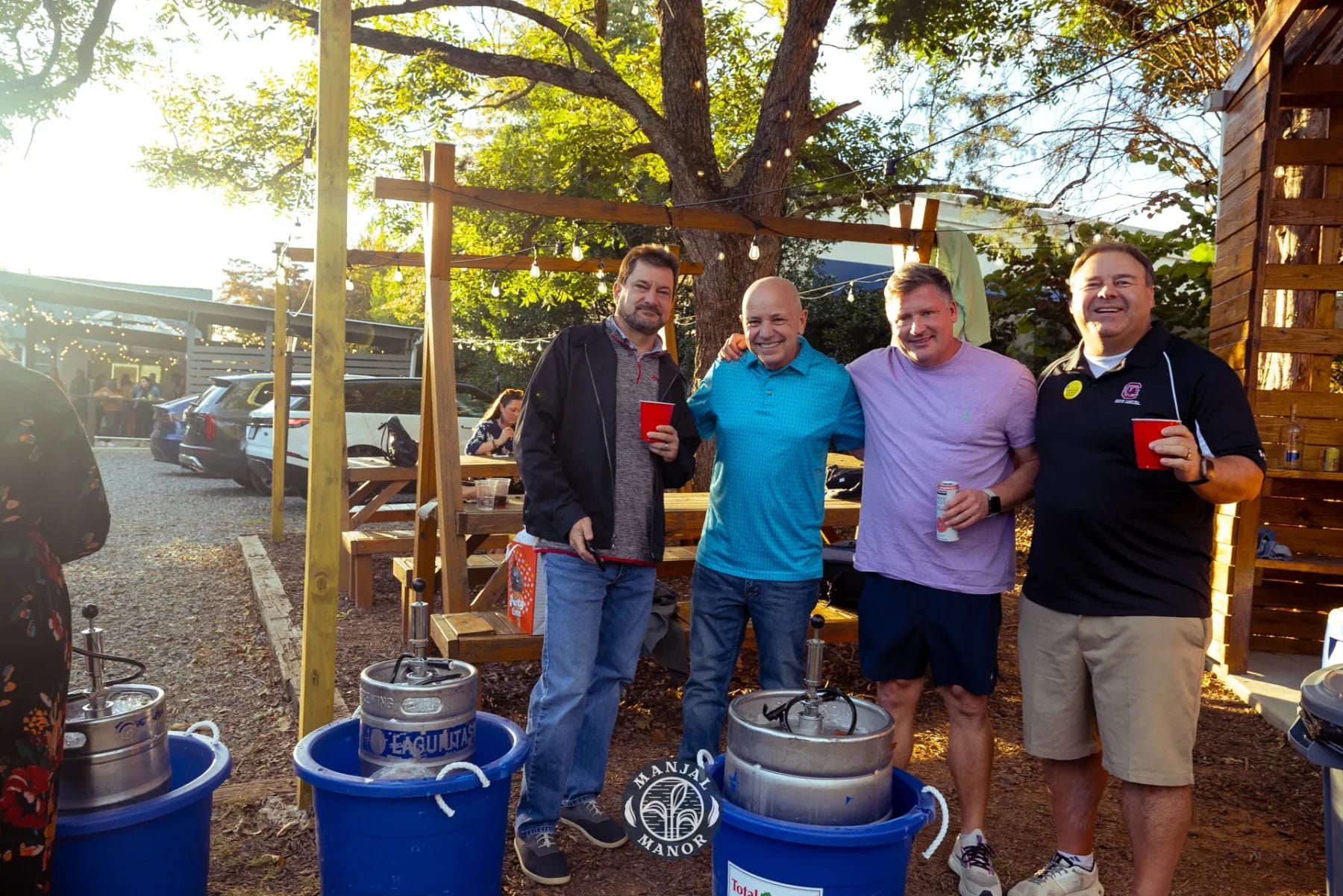 Four men stand outdoors holding drinks, smiling at the camera. They are next to barrels and coolers under a wooden structure, with trees and sunlight in the background. Casual and festive atmosphere. Huntersville event venue