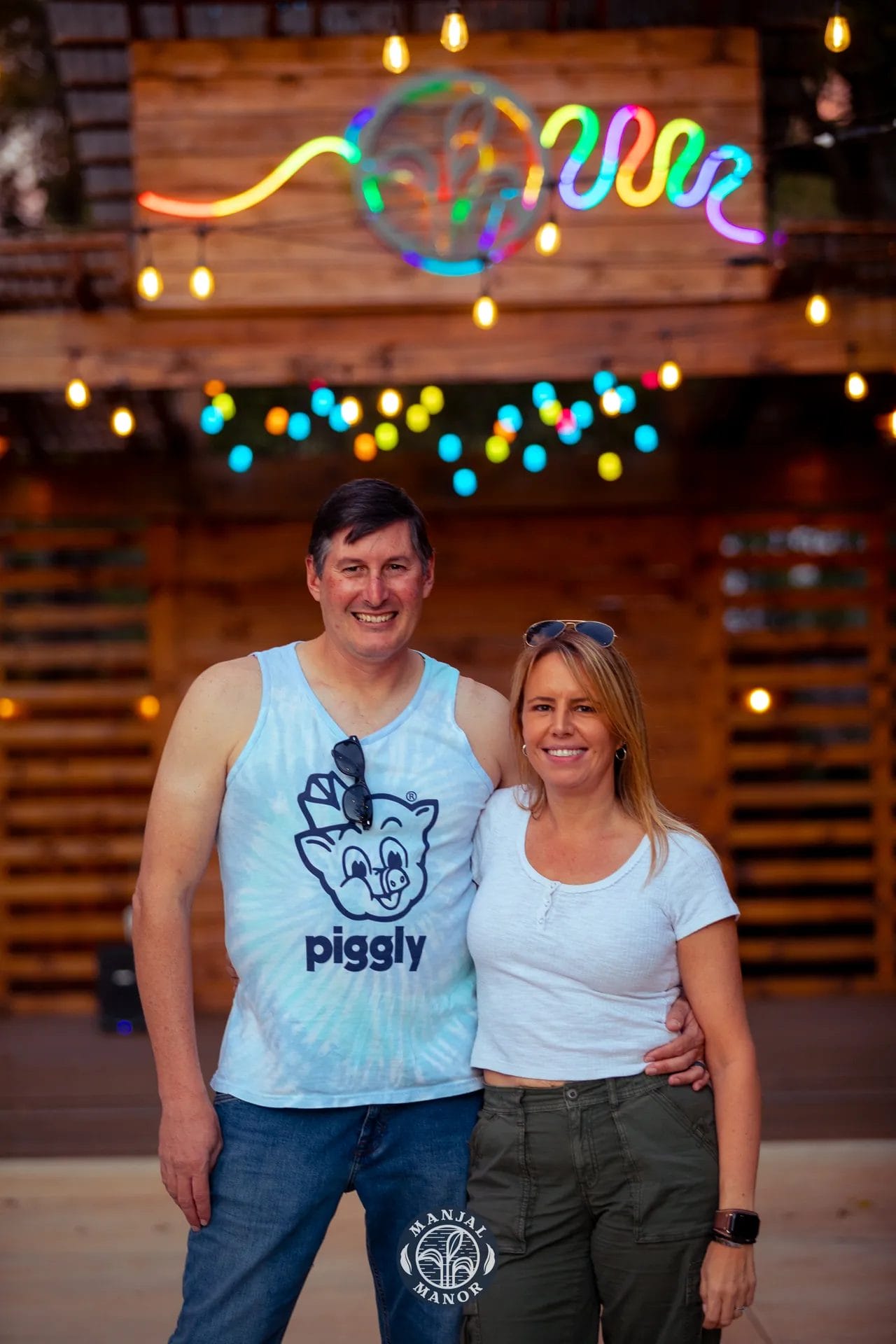 A smiling couple poses in front of a rustic wooden backdrop with a colorful neon sign above them. The man wears a blue tie-dye tank top with a cartoon pig design, while the woman is in a white shirt. String lights decorate the scene. Huntersville event venue