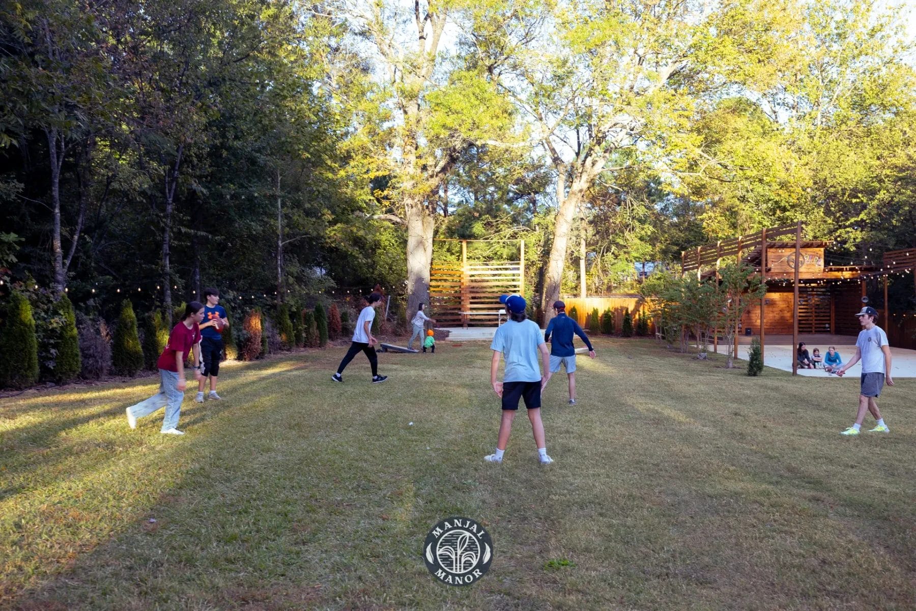 A group of people playing a casual soccer game on a grassy field surrounded by trees and wooden structures. It is a sunny day, and the participants are dressed in casual sportswear. Huntersville event venue