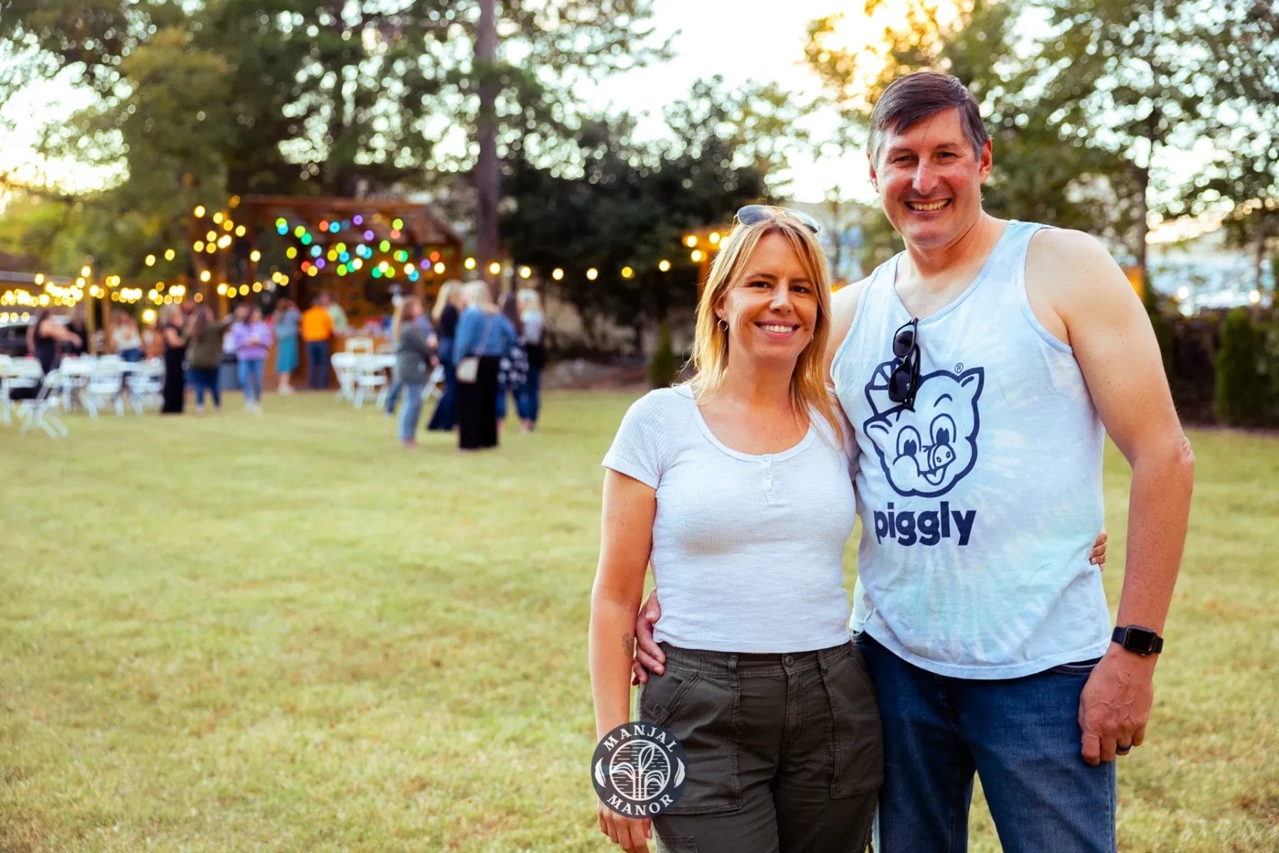 A smiling couple stands together on a grassy area during an outdoor event. The man is wearing a sleeveless shirt and the woman is in a white top. Festive lights and people are visible in the background under trees at sunset. Huntersville event venue