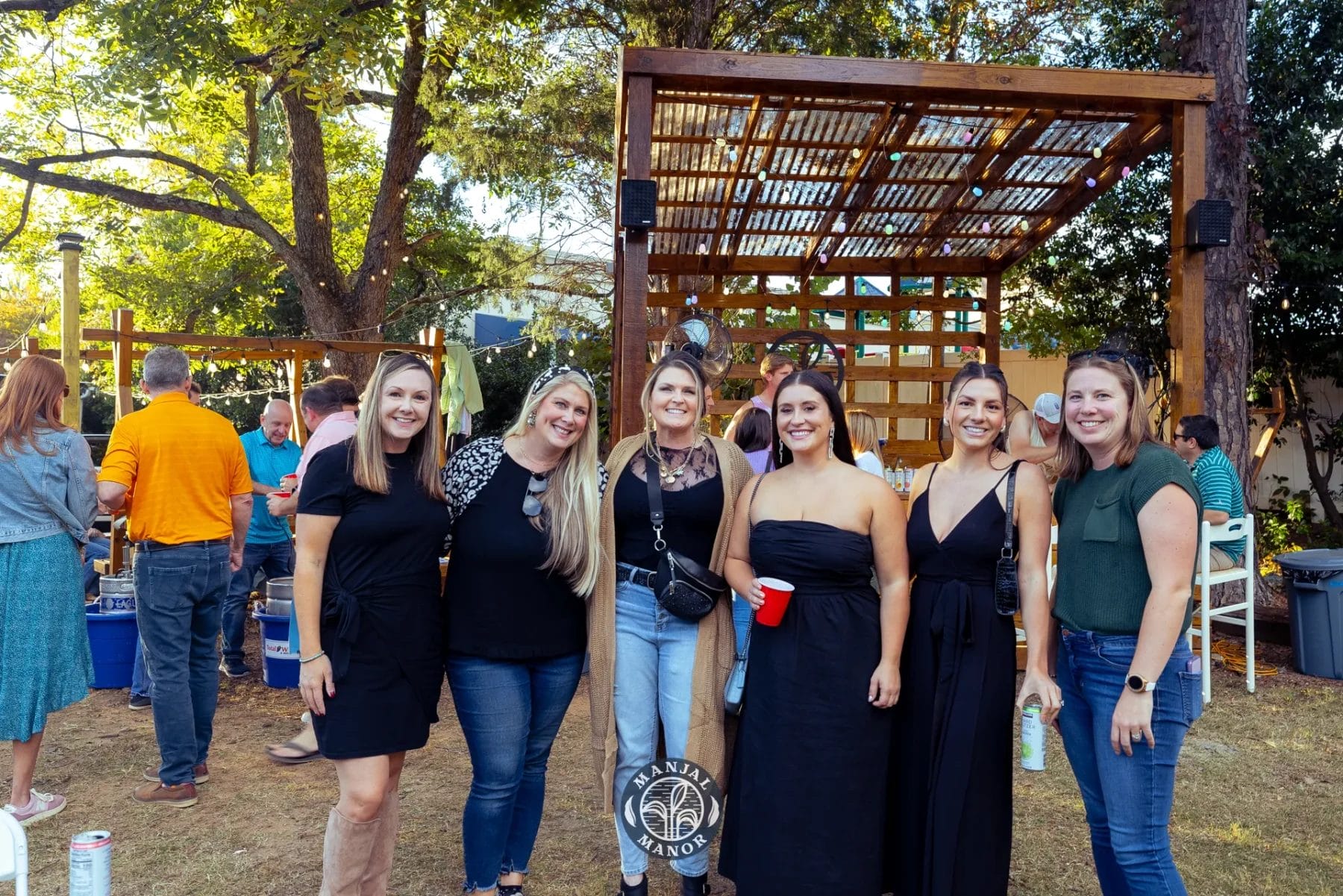 A group of six women standing together smiling outdoors. They're in a garden area with wooden structures and trees. Some hold drinks in their hands. The sunlight creates a warm, cheerful atmosphere. Huntersville event venue