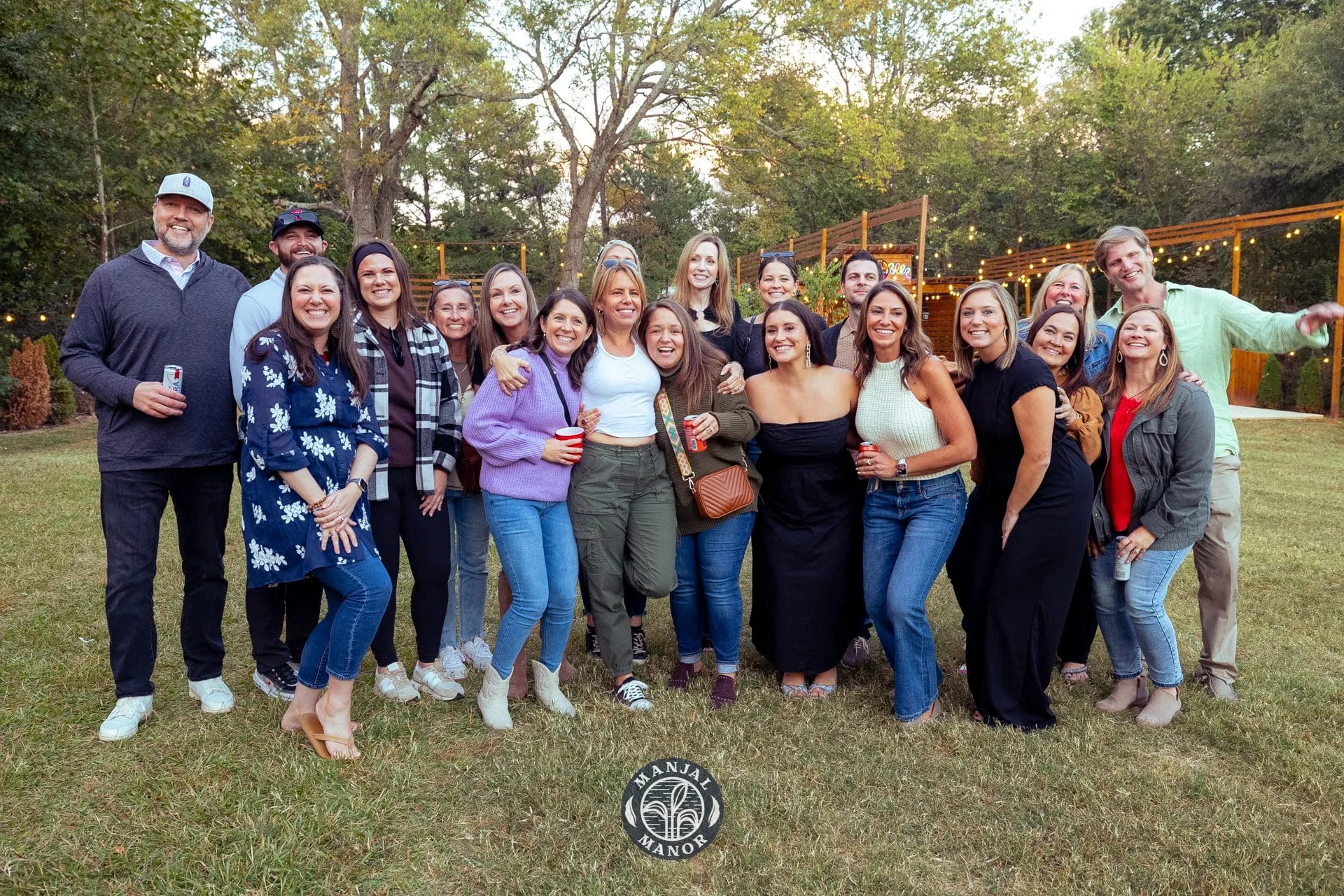 A group of people smiling and posing for a photo outdoors in a grassy area with trees in the background. They are holding drinks and appear to be enjoying a casual gathering. The scene is well-lit with string lights adding a warm ambiance. Huntersville event venue