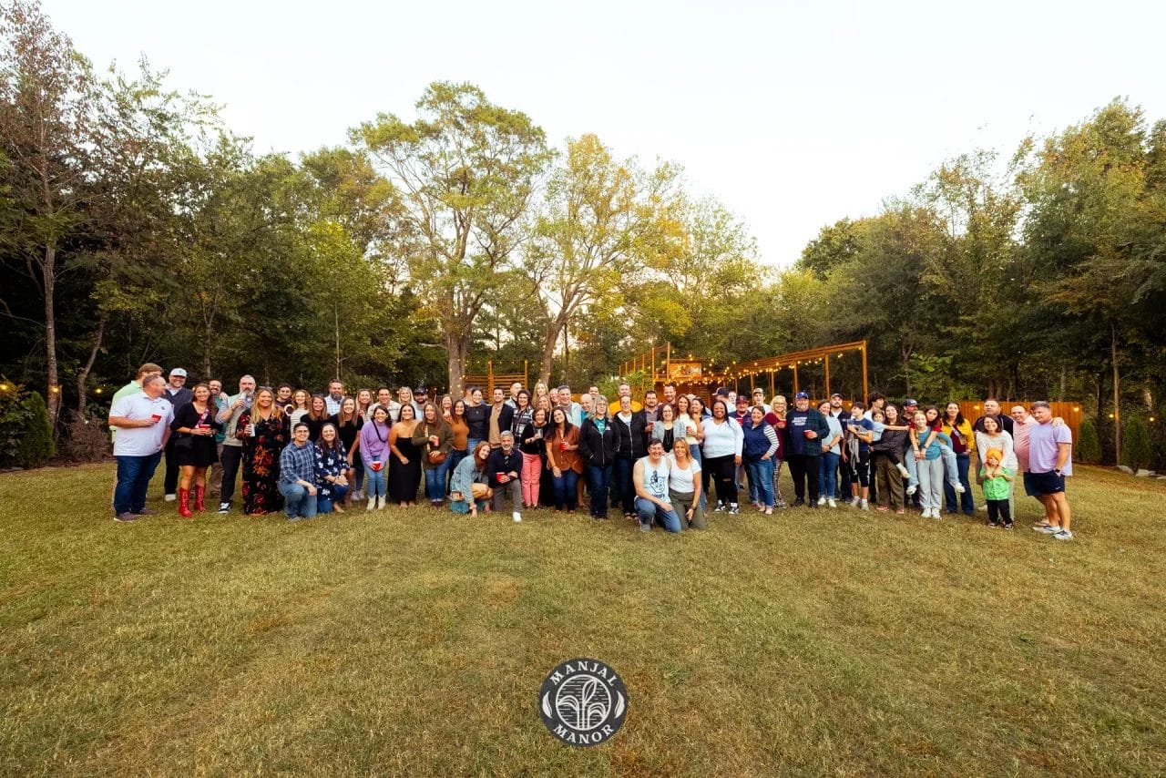 A large group of people posing outdoors on a grassy area with trees in the background. Some are standing, while others kneel or sit. There are festoon lights hanging among the trees, and a logo on the grass says 