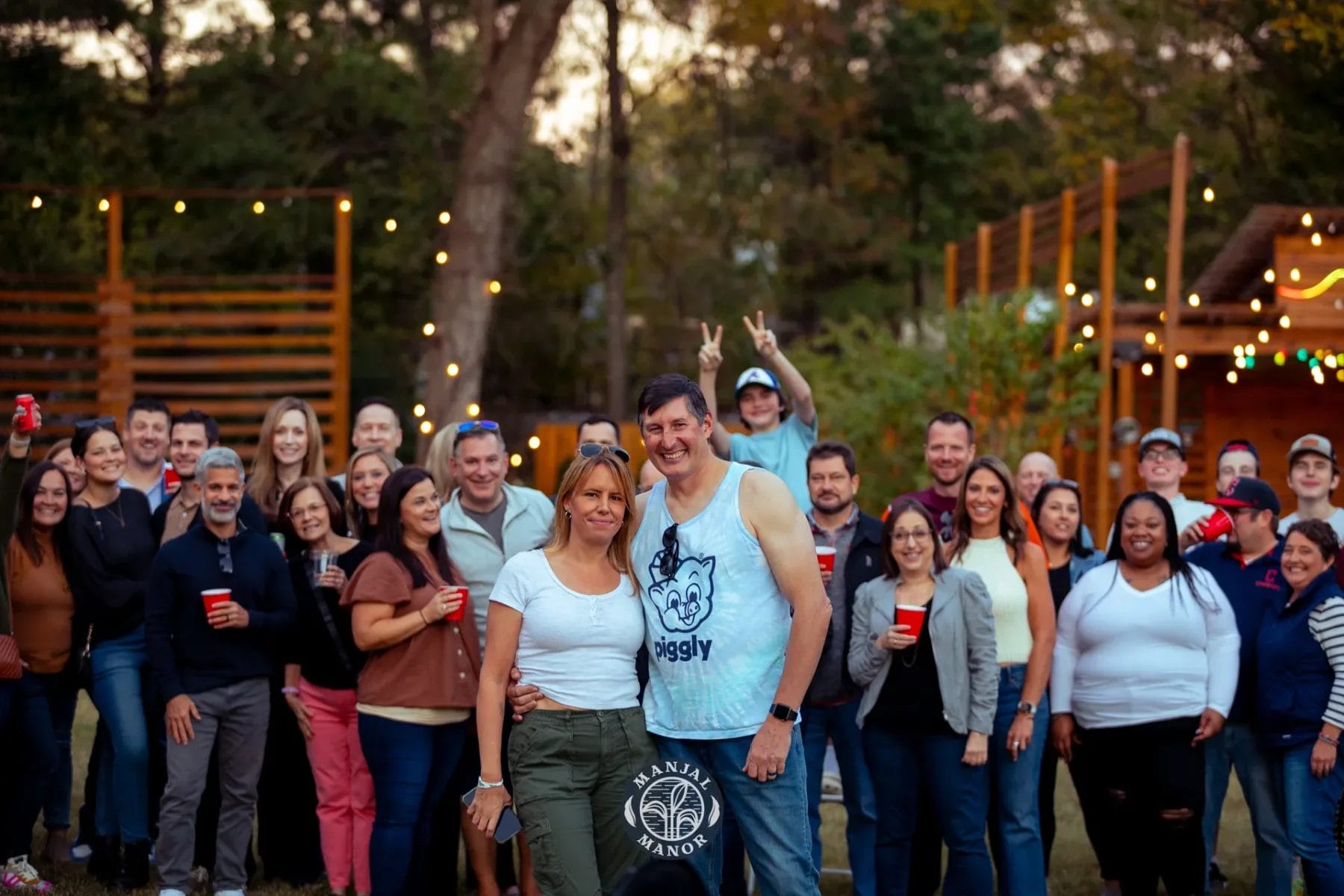 A large group of people pose together outdoors, smiling and holding drinks. They stand in a grassy area with trees and string lights in the background. A man and woman stand at the front, with the logo "Janja Manor" on the image. Huntersville event venue