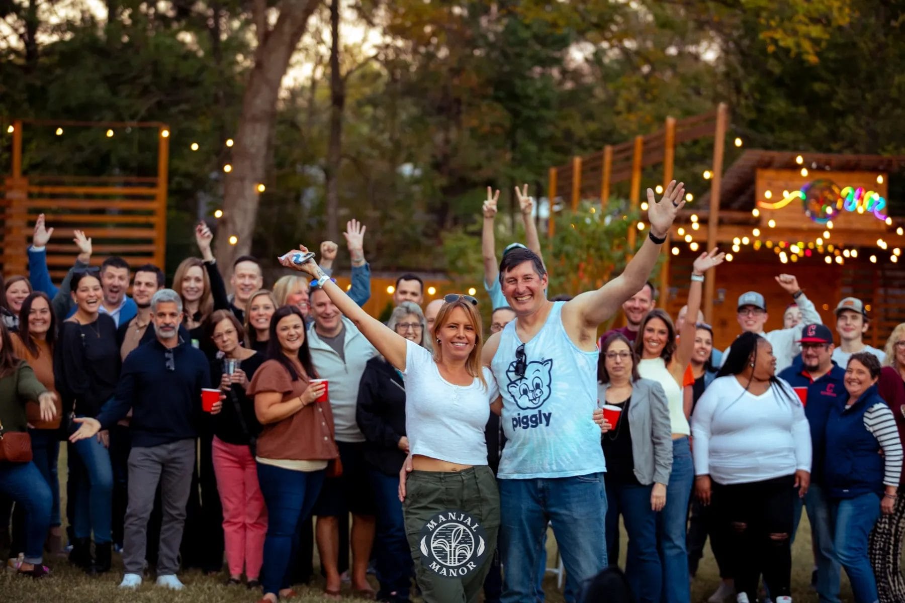 A large group of people smiling and cheering outdoors. Two people stand at the front, posing with their arms raised. String lights and trees are in the background, creating a festive atmosphere. Huntersville event venue